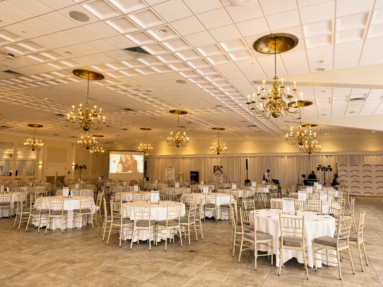 Bright ballroom set for a nonprofit gala with round tables, white linens, gold chairs, chandeliers and a branded step and repeat backdrop